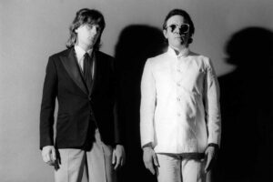 Two men stand against a plain background; one wears a dark blazer and tie with light pants, while the other wears a buttoned white suit and large round sunglasses. Both look forward with neutral expressions.