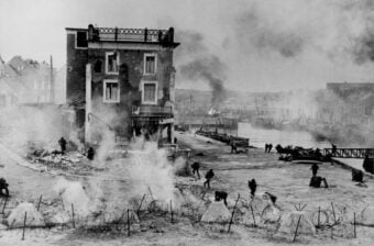 Black and white photo of soldiers advancing through smoke and barbed wire near a damaged building by a waterfront during a battle, with more smoke and destruction visible in the background.