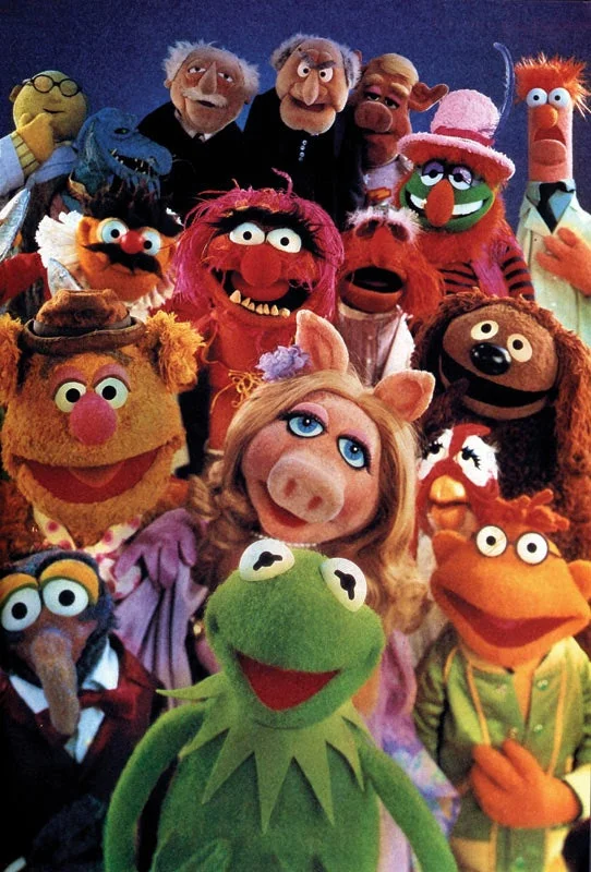 A group of colorful Muppets, including Kermit the Frog, Miss Piggy, Fozzie Bear, Gonzo, and others, smile and pose together against a blue background.