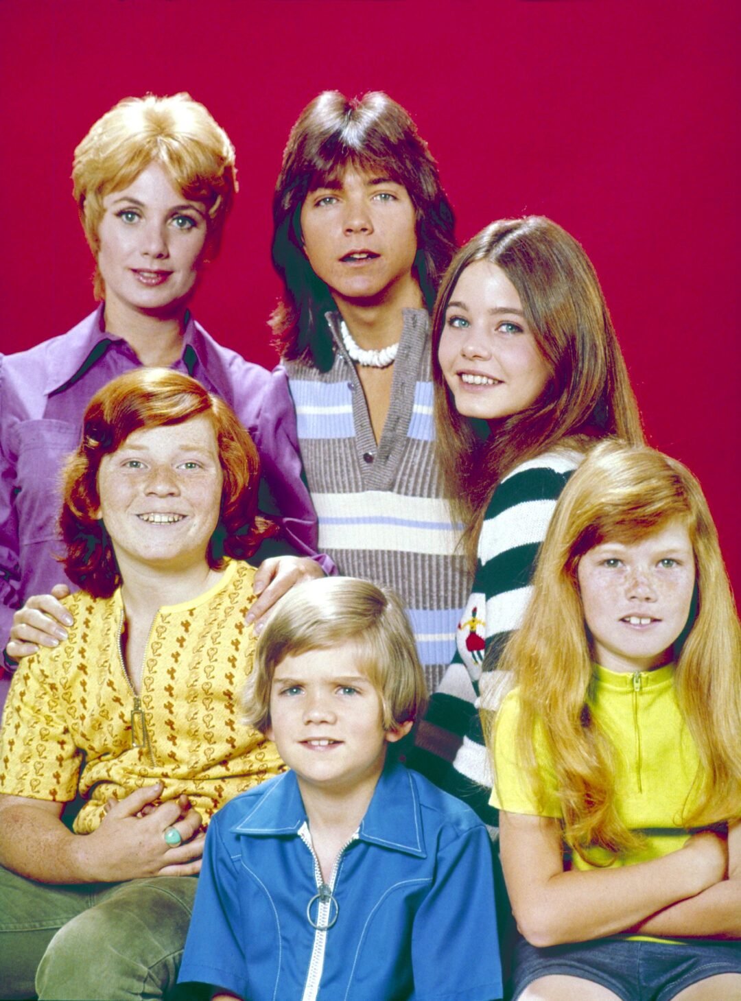 A group of six people, three adults and three children, pose together smiling in front of a solid red background. They are dressed in colorful, casual 1970s-style clothing.