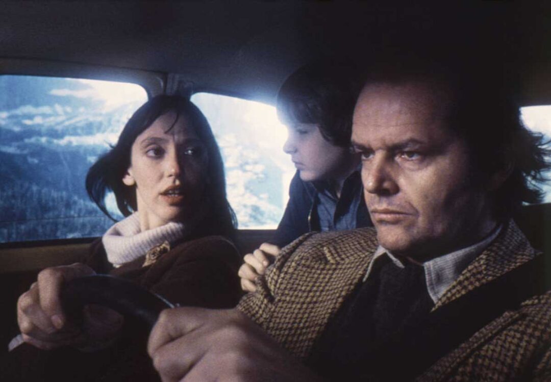 A woman is driving a car while looking at a serious man in the passenger seat; a young boy is sitting between them, gazing at the man. Snowy mountains are visible through the car window.