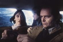 A woman is driving a car while looking at a serious man in the passenger seat; a young boy is sitting between them, gazing at the man. Snowy mountains are visible through the car window.