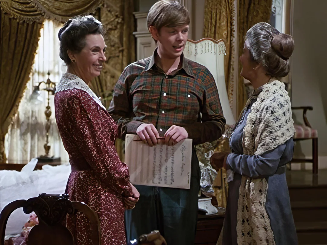 A young man holding a sheet of paper stands between two older women in a Victorian-style room, all smiling and engaged in conversation. The room features elegant curtains and antique furniture.