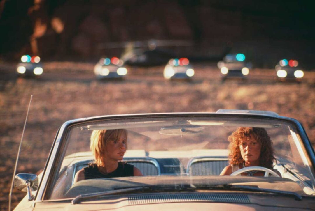 Two women sit in the front seats of a convertible car in a desert setting, looking tense. Behind them, several police cars with flashing lights and a helicopter are visible in the background.