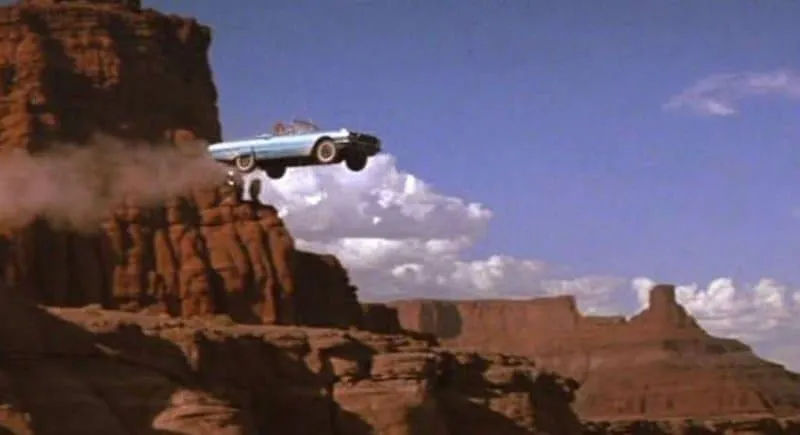 A blue convertible car soars off a cliff edge in a desert canyon landscape under a blue sky with scattered clouds.