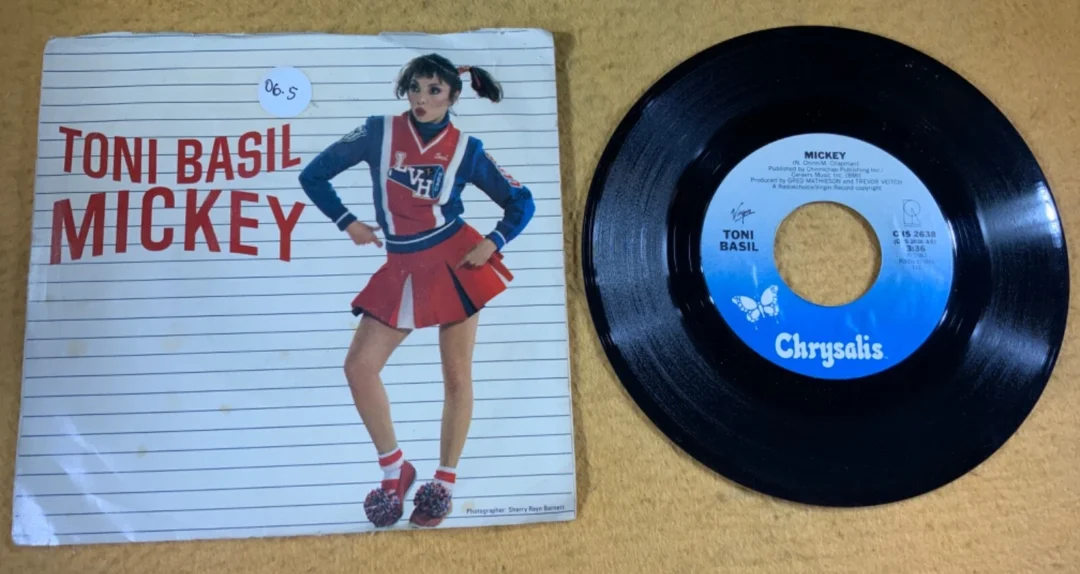 A vinyl record of Toni Basil’s single “Mickey” sits next to its cover, which shows Toni Basil dressed as a cheerleader, standing in a confident pose against a lined background with bold red text.