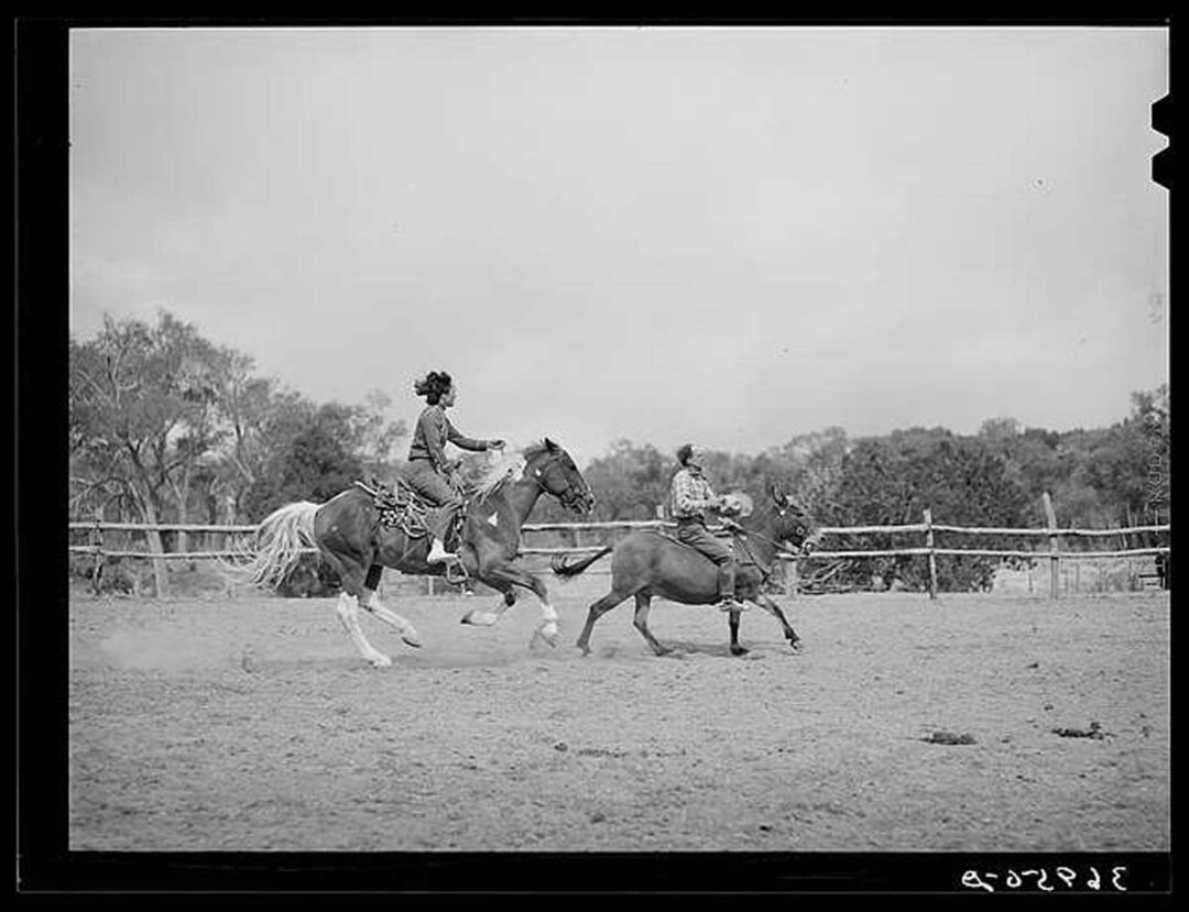 Two people on horseback practice roping in a dusty corral, with trees and a wooden fence in the background under a cloudy sky.