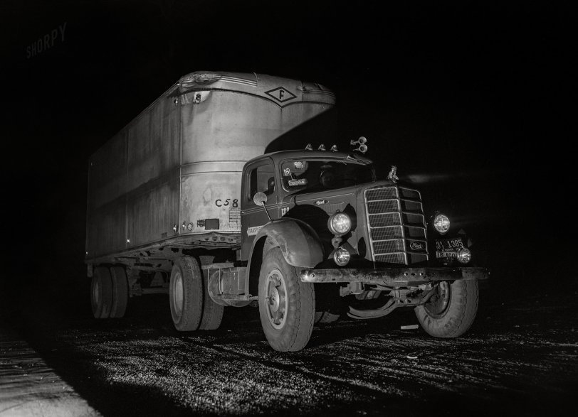 Black and white photo of a vintage semi-truck and trailer parked on a dark road at night, with illuminated headlights and distinctive grille details visible.