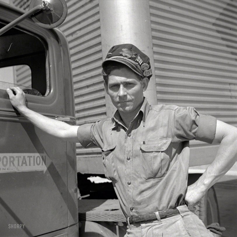 A man in a short-sleeve work shirt and cap stands with one arm resting on a truck door, looking at the camera. A large industrial building is in the background.