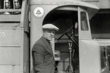 A man in a flat cap and pinstripe suit leans against a vintage delivery truck, looking toward the camera. The truck&rsquo;s door is open, revealing part of the interior and a mounted milk bottle crate.