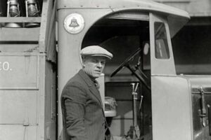 A man in a flat cap and pinstripe suit leans against a vintage delivery truck, looking toward the camera. The truck’s door is open, revealing part of the interior and a mounted milk bottle crate.