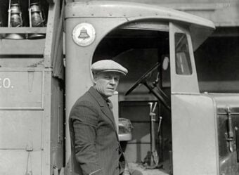 A man in a flat cap and pinstripe suit leans against a vintage delivery truck, looking toward the camera. The truck&rsquo;s door is open, revealing part of the interior and a mounted milk bottle crate.