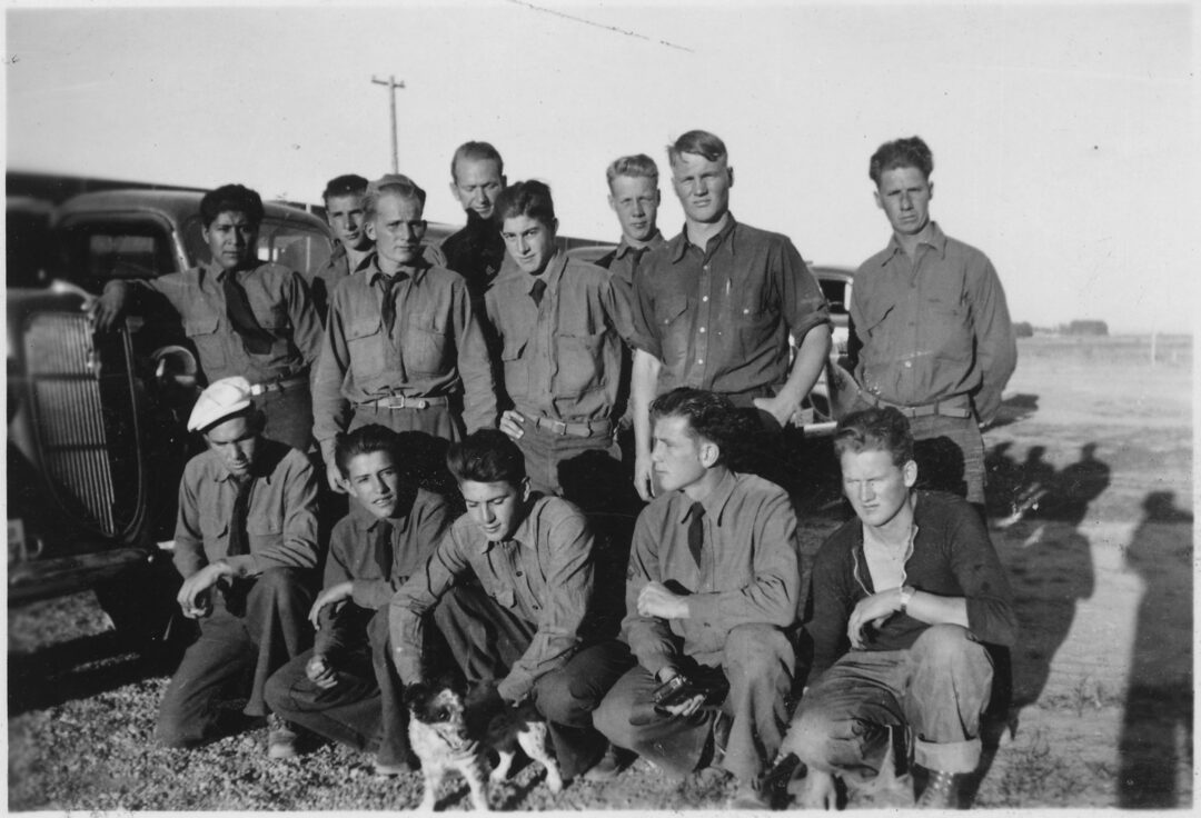 A black-and-white photo of thirteen young men wearing similar uniforms, some standing and some squatting outside on a sunny day, with a car and a small dog in the foreground. The background shows open land and sparse objects.