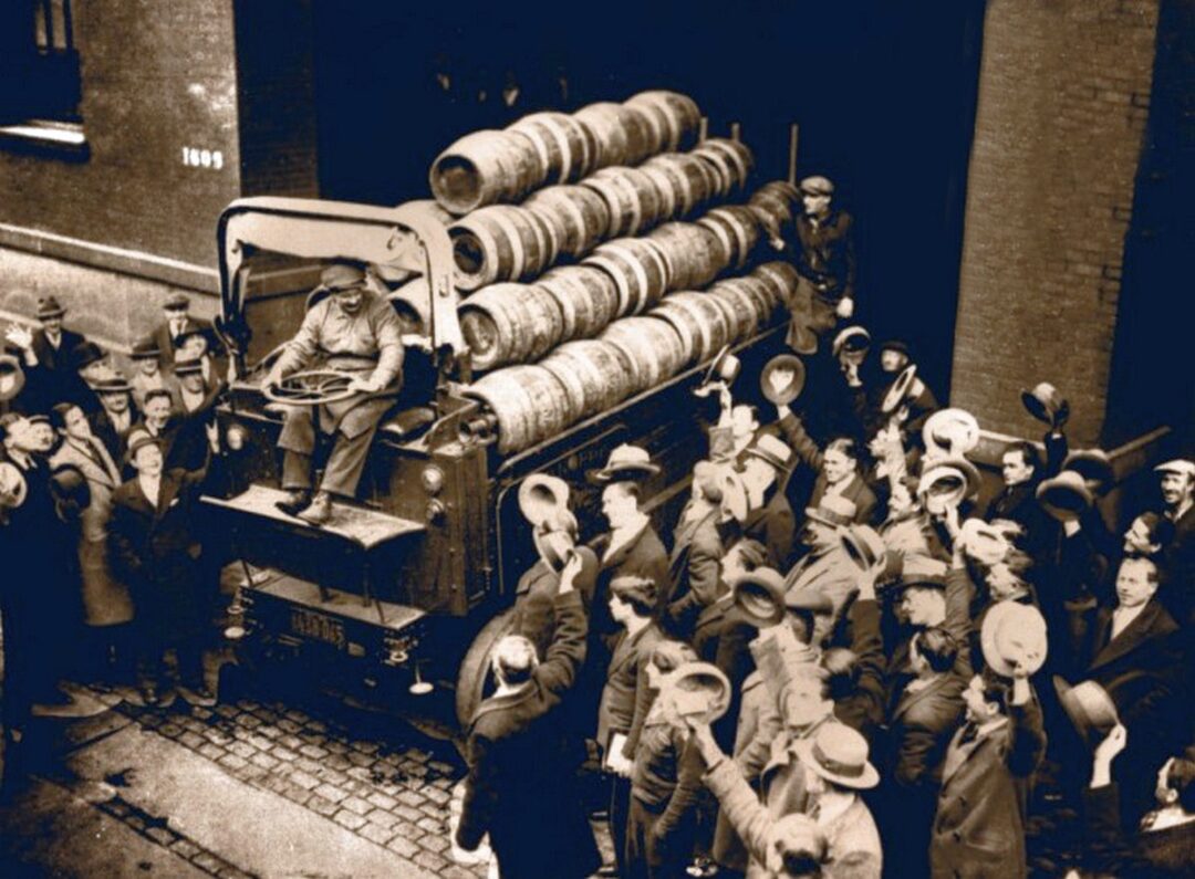 A crowd of people in old-fashioned clothing gathers around a truck loaded with barrels, cheering as a driver unloads them in front of a brick building. The photo appears to be from the early 20th century.