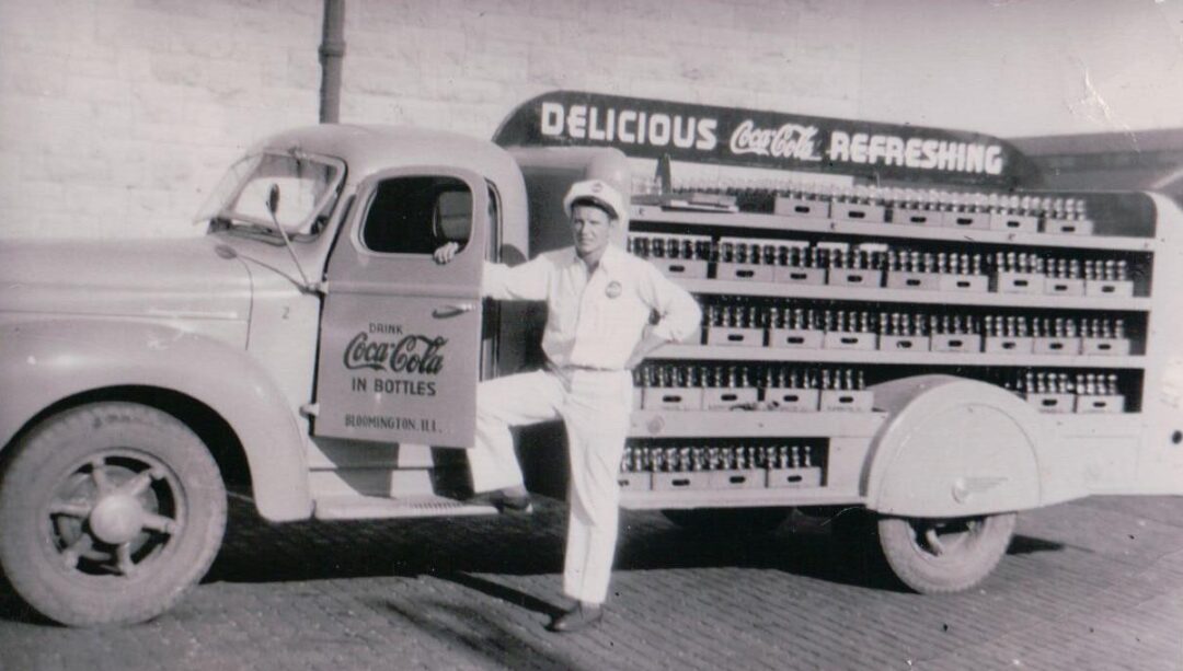 A vintage black-and-white photo of a man in a uniform standing beside a classic Coca-Cola delivery truck loaded with glass soda bottles. The truck displays "Delicious Coca-Cola Refreshing" on its sign.