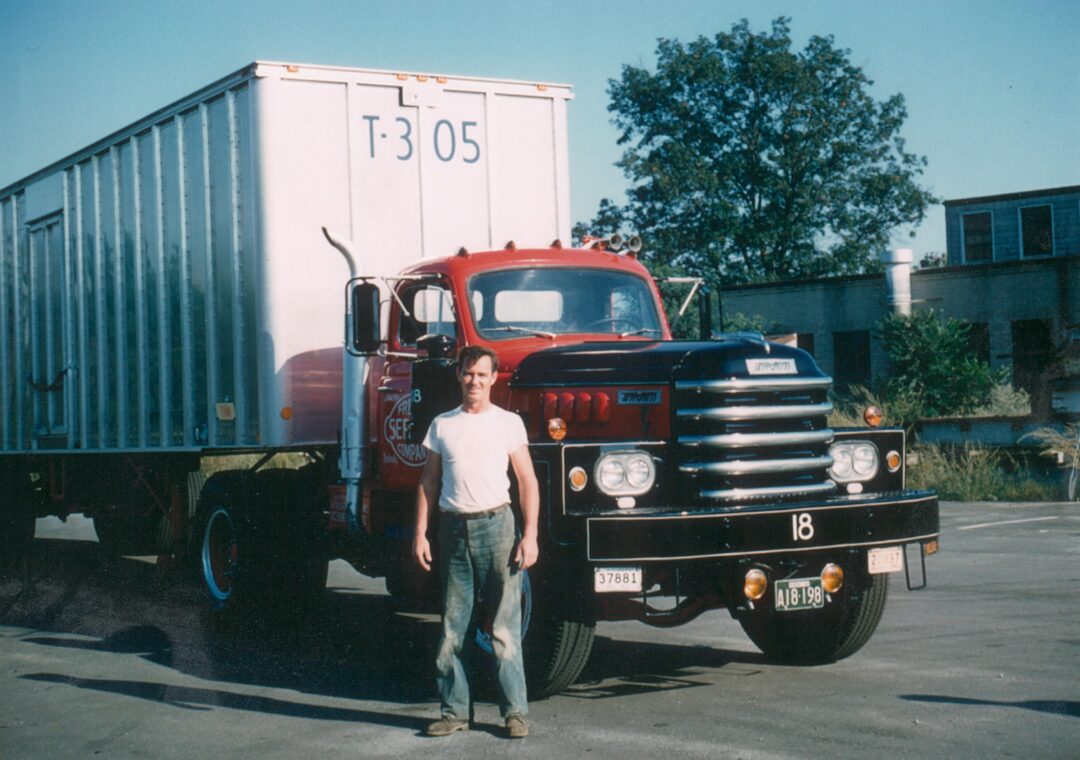 A man in a white shirt and jeans stands in front of a vintage red and black semi-truck with a large white trailer, parked on a paved lot with trees and a building in the background.