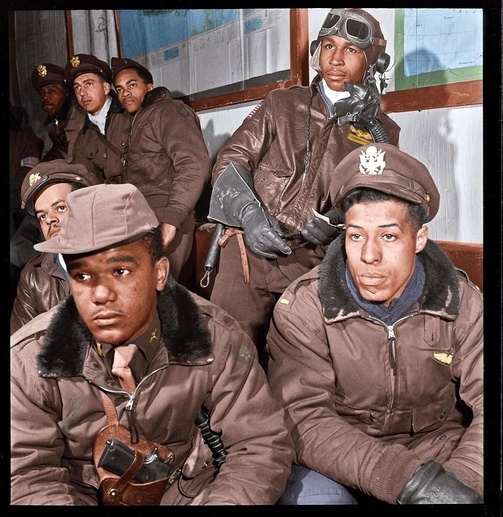 A group of Tuskegee Airmen in brown military uniforms and caps sit together indoors, some looking focused, one wearing goggles and gloves, with maps on the wall behind them.