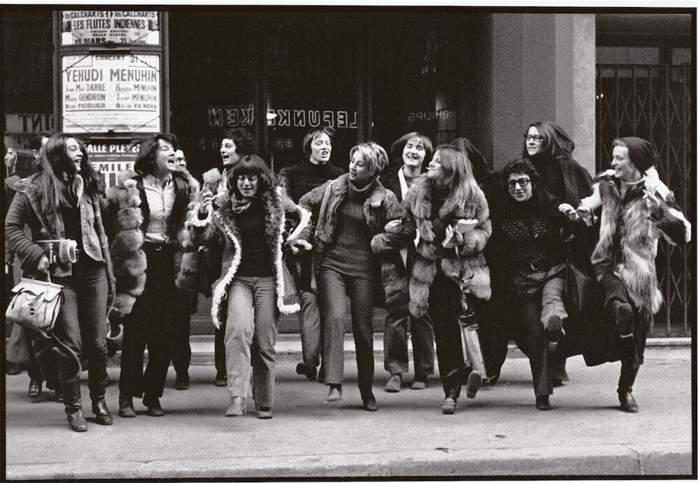 A group of women, dressed in winter clothing, hold hands and smile as they march energetically down a city street. A sign with concert information is visible in the background.