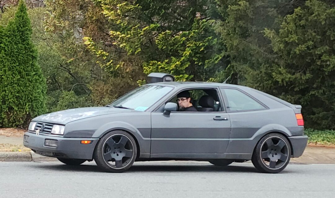 A person wearing glasses sits in the driver's seat of a gray two-door hatchback car parked on a street, with trees and greenery in the background.