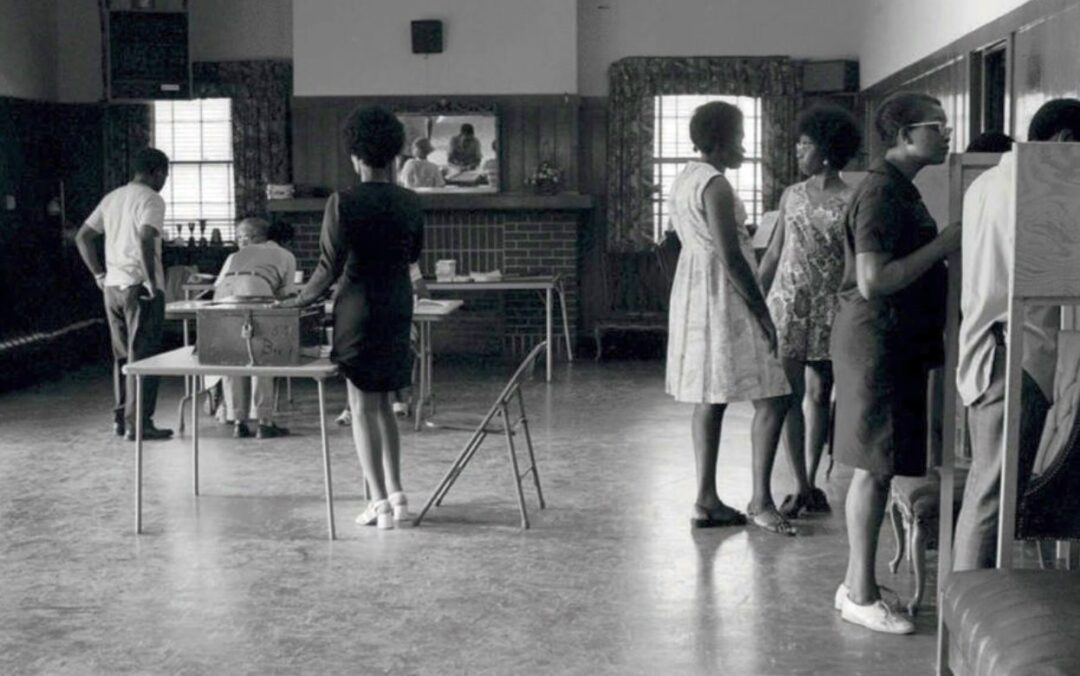 Black-and-white photo of people inside a polling place. Some stand in line or vote at booths, while others sit at tables checking in voters. A mirror and fireplace are visible in the background.