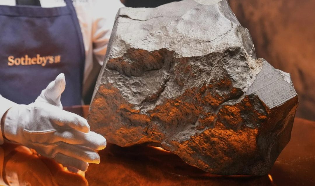 A person wearing white gloves and a “Sotheby’s” apron presents a large, irregularly shaped gray rock, possibly a meteorite, displayed on a shiny surface with warm lighting.