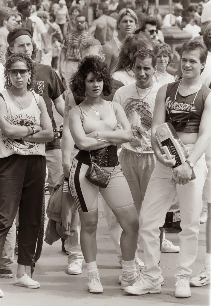 A group of young adults in casual 1980s athletic wear stand outdoors in a crowd. One person holds a boombox, and another has arms crossed. The scene is lively and captures retro street fashion.