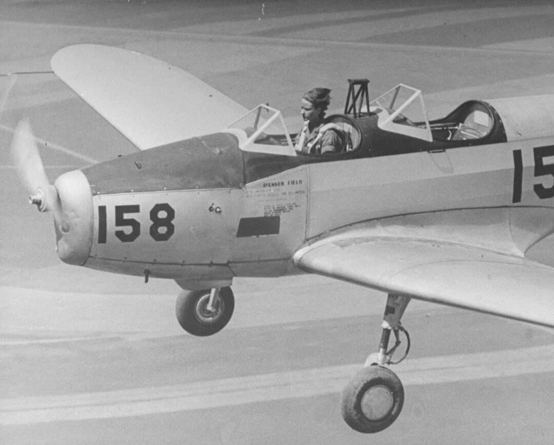 A woman pilot flies a small single-engine airplane marked with the number 158. The cockpit is open, and the plane is shown in flight with the landing gear extended.