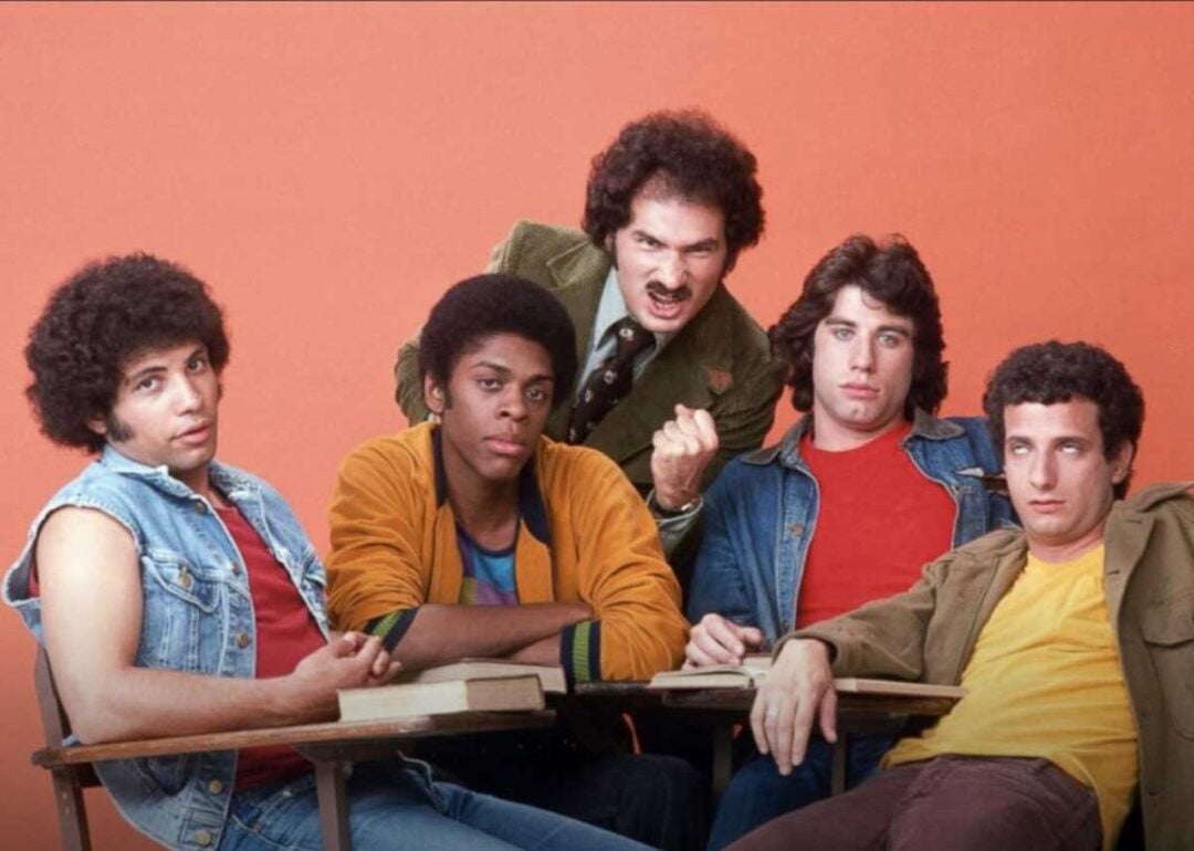 Five young men sit and lean around school desks against an orange backdrop, looking at the camera with various expressions. The man in the back stands, fist raised, while the others sit in casual poses.