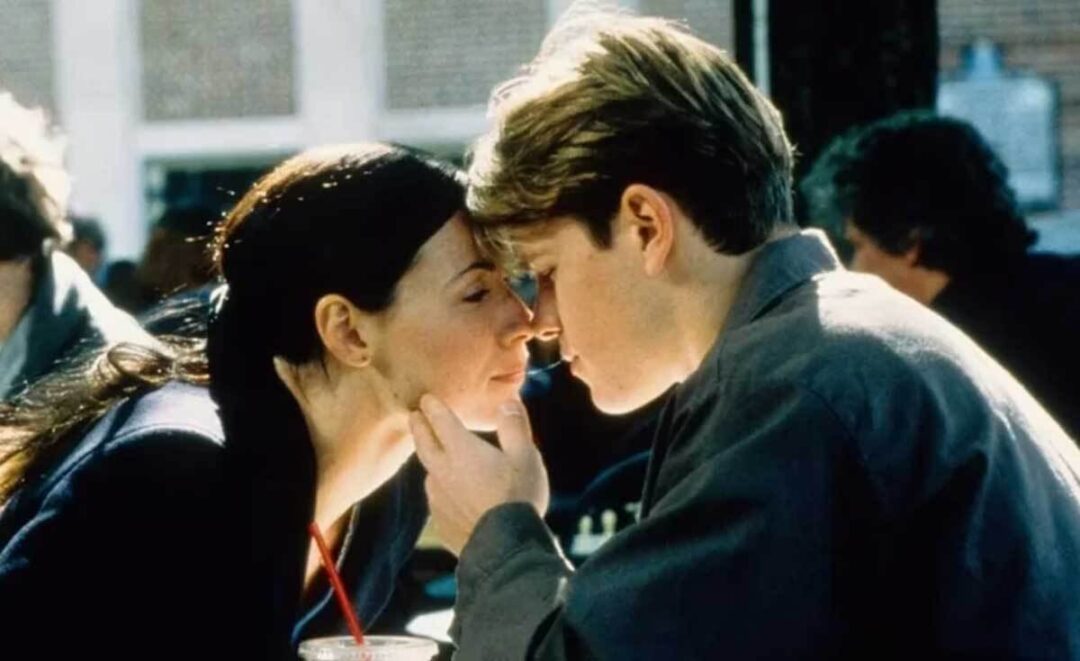 A woman and a man sit closely at an outdoor café, touching foreheads and gazing into each other’s eyes tenderly. The man gently holds the woman’s chin, and a drink with a red straw is visible in the foreground.