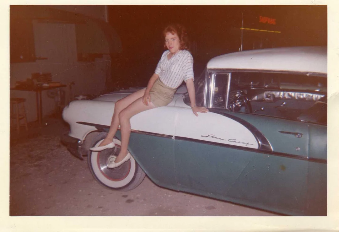A young woman in a plaid shirt and shorts sits on the front fender of a vintage green and white car at night. The car is a Pontiac Star Chief, and the scene appears casual and relaxed.