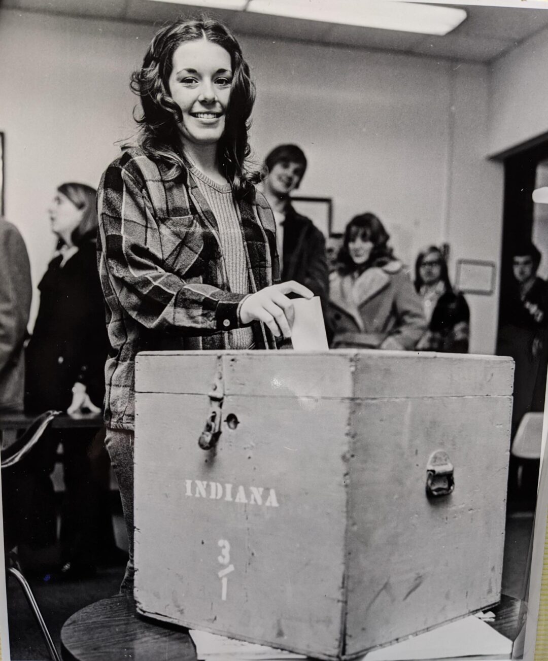 A young woman smiles as she casts her ballot into a box labeled "Indiana," while others wait in line behind her in a polling place. The photo is black and white.