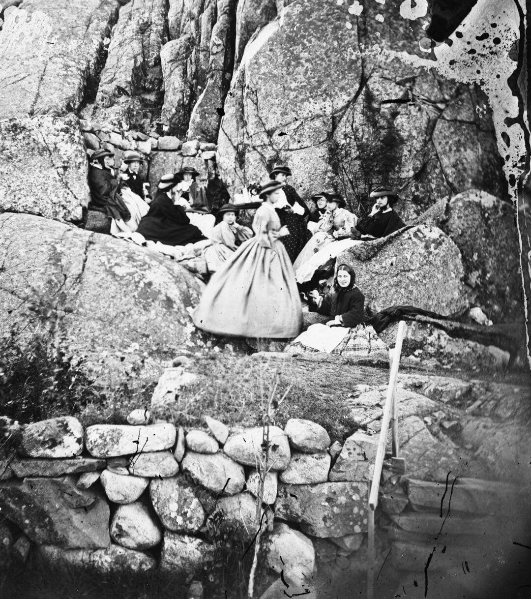 A group of people in 19th-century clothing sit and recline on rocky terrain, surrounded by large boulders and stone walls; one woman stands in the foreground wearing a full skirt. The scene is outdoors.