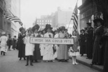 A group of women and girls in early 1900s clothing stand on a city street, holding American flags and a banner that reads &ldquo;I WISH MA COULD VOTE,&rdquo; advocating for women&rsquo;s suffrage.