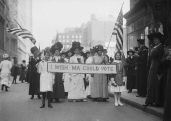 A group of women and girls in early 1900s clothing stand on a city street, holding American flags and a banner that reads &ldquo;I WISH MA COULD VOTE,&rdquo; advocating for women&rsquo;s suffrage.