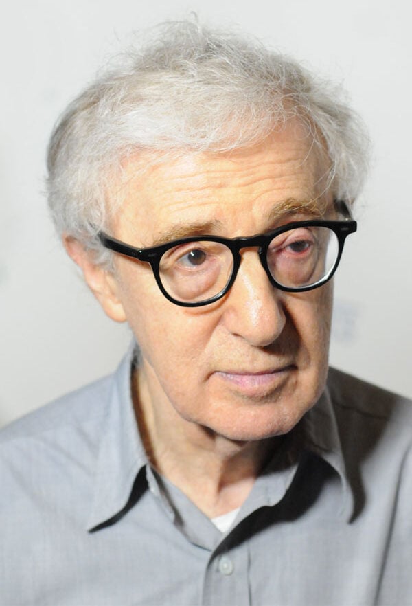 An older man with light gray hair and black-rimmed glasses is looking slightly to the side. He is wearing a light gray button-up shirt against a plain, light background.
