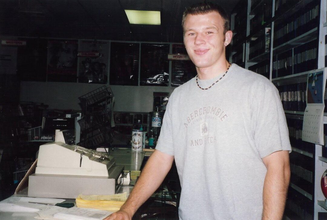 A young man wearing a gray Abercrombie t-shirt and a beaded necklace stands behind a counter with a cash register and paperwork in a video rental store. Shelves of tapes and movie posters are visible in the background.