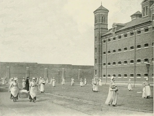 Black-and-white photo of women in long dresses and aprons walking or carrying laundry in the yard of a large, brick institutional building, possibly a prison or workhouse, with high walls and watchtowers.