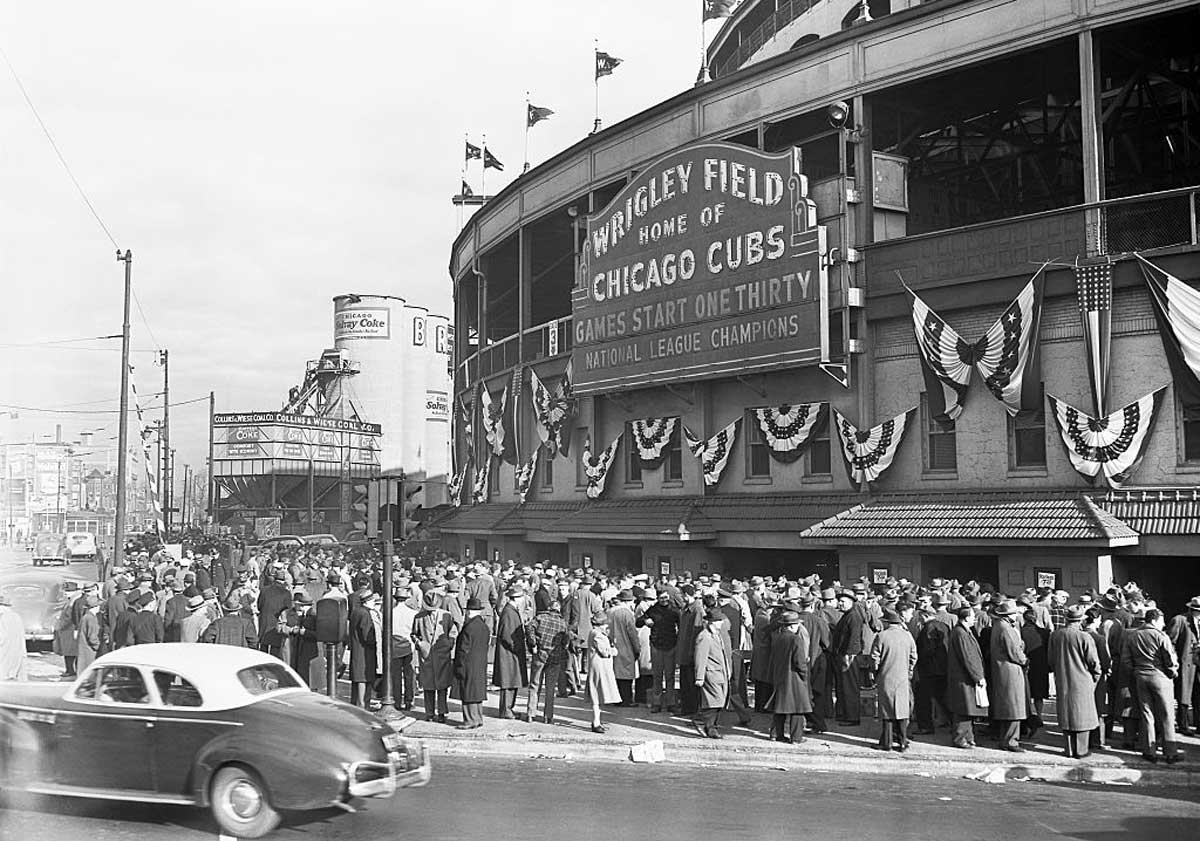 A large crowd gathers outside Wrigley Field in Chicago, decorated with bunting. People in coats and hats wait near the entrance beside a classic car, under a sign for the 1945 National League Champion Chicago Cubs.
