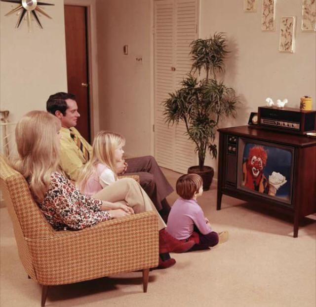 A family of four sits in a living room watching TV. The show features a clown holding a hamburger. The room has a patterned sofa, potted plant, and mid-century decor, suggesting a retro 1970s setting.