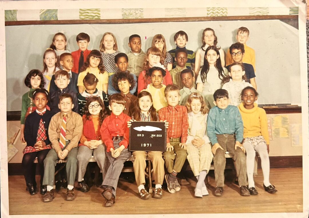 A diverse elementary school class from 1971 poses for a group photo; children are seated and standing in rows, with one child in front holding a sign that reads "GNA GR2 1971." The background features classroom decorations.