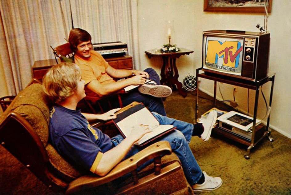Two people sit in a retro living room watching MTV on a vintage television. One person holds a notebook, both are smiling and relaxed. The room features brown furniture, green carpet, and a shelf with electronics.