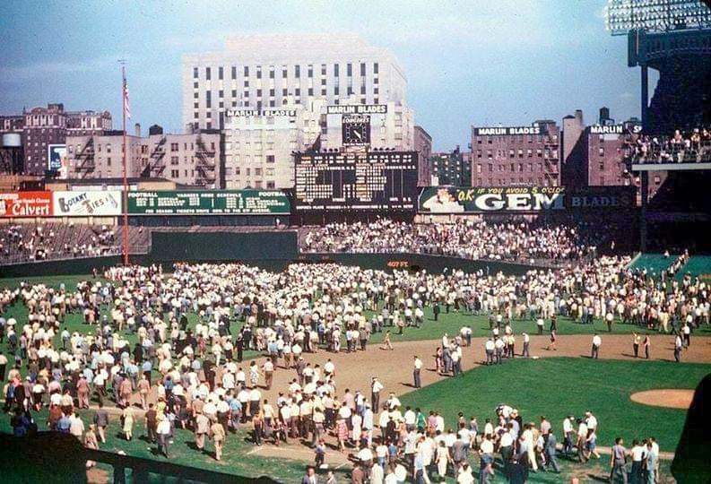 A large crowd of people fills a baseball field and outfield on a sunny day, with scoreboards, advertisements, and city buildings visible in the background. The stands are full, suggesting a significant event has just occurred.