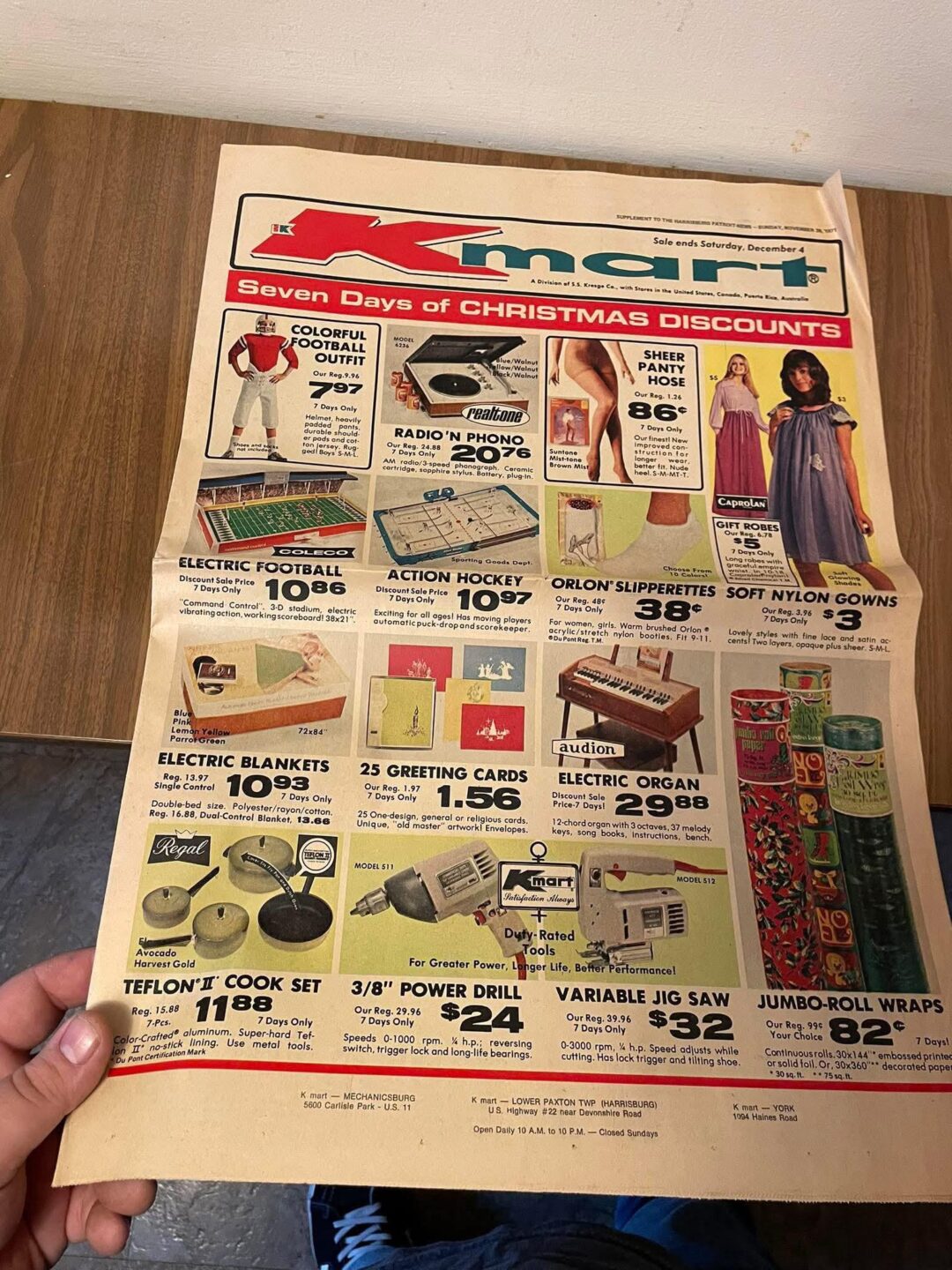 A person holds a vintage Zayre store flyer advertising "Seven Days of Christmas Discounts" on toys, clothing, kitchenware, and gifts, with colorful images and prices for each item on a wooden table.