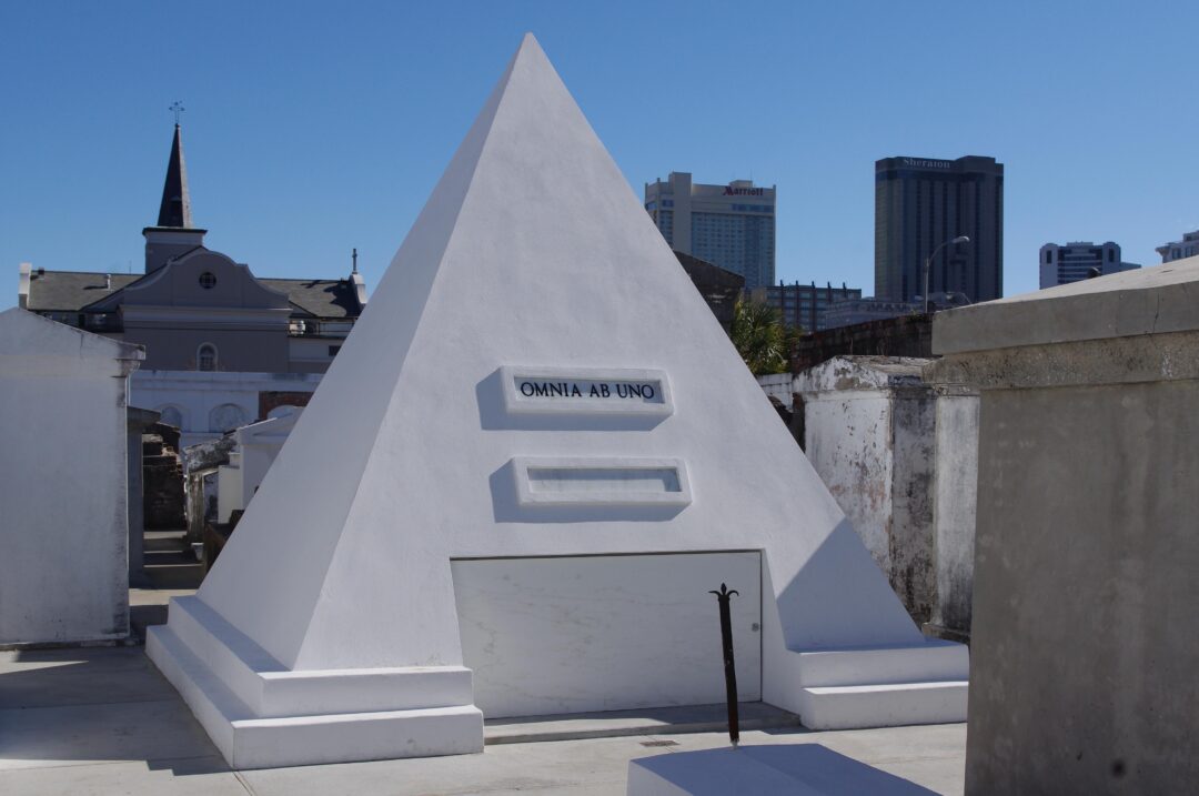 A white pyramid-shaped tomb with "Omnia Ab Uno" written on it stands in a cemetery, surrounded by other tombs and city buildings, under a clear blue sky.