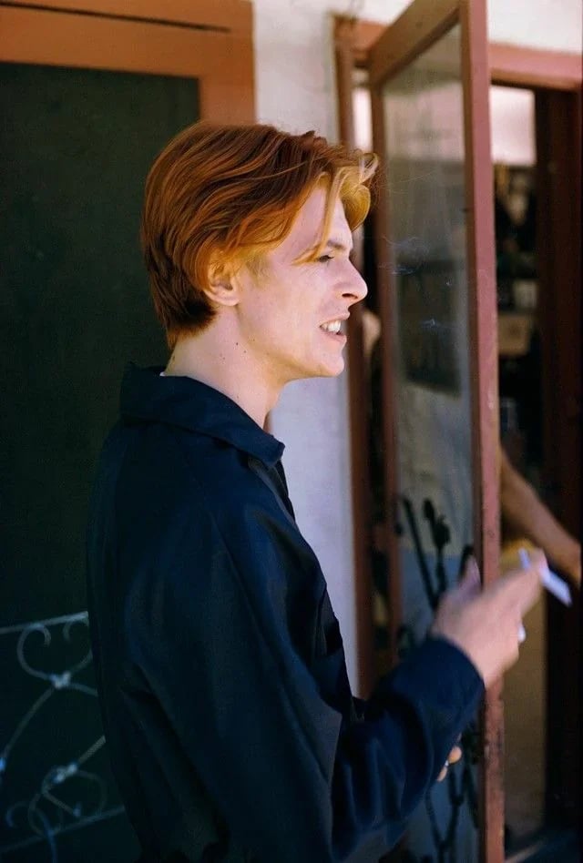 A person with short red hair, wearing a dark shirt, stands outside near a doorway, holding a cigarette and looking off to the side, with a slight smile on their face.