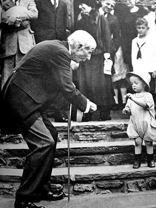 An elderly man with a cane bends down to interact with a small child standing on stone steps, while a group of people in early 20th-century clothing watch in the background.
