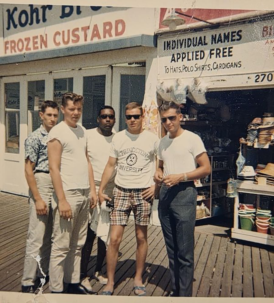 Five young men stand together on a wooden boardwalk in front of a frozen custard shop and a store selling hats and shirts. They wear casual summer clothes, and it appears to be a sunny day in the 1960s.