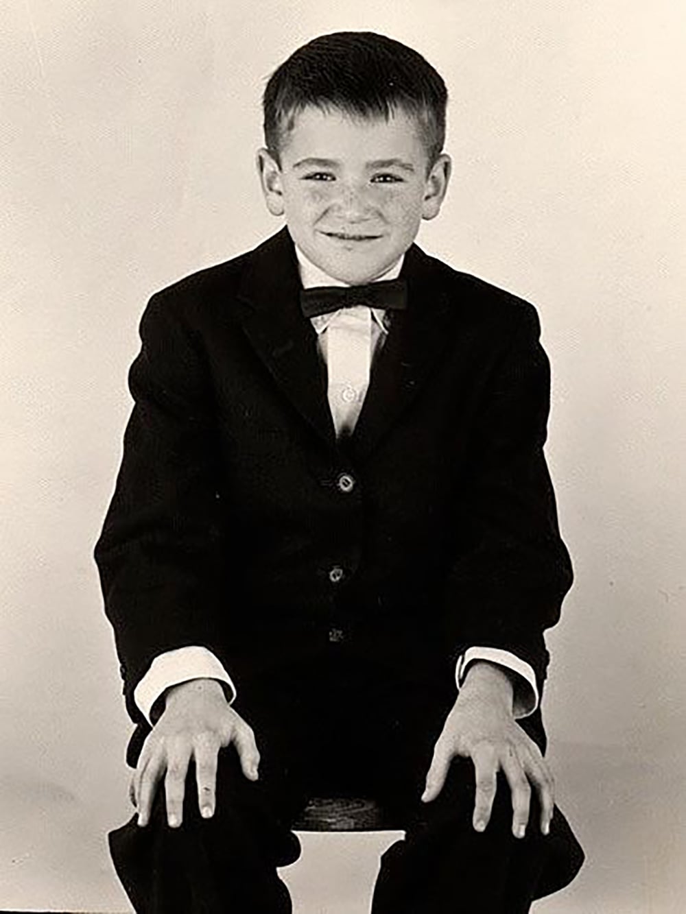 A young boy wearing a dark suit, white shirt, and bow tie sits on a stool with his hands on his knees, smiling at the camera against a plain background.