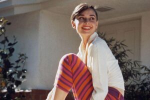 A young woman with short dark hair sits barefoot on a bench outdoors, smiling and looking to the side. She wears a white long-sleeve top and pink-and-orange striped pants. A house and greenery are visible in the background.