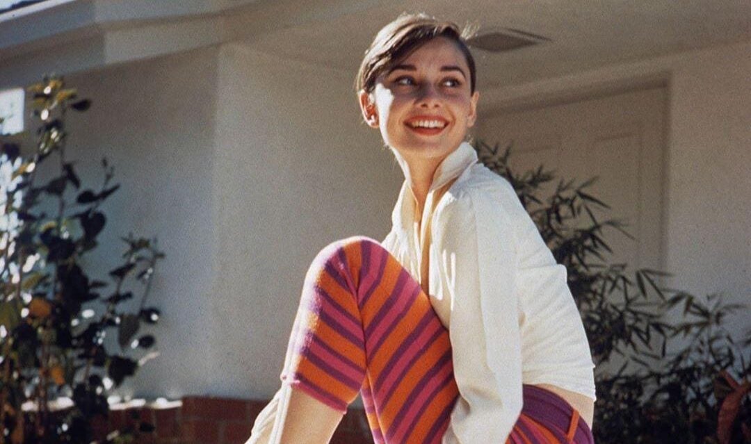 A young woman with short dark hair sits barefoot on a bench outdoors, smiling and looking to the side. She wears a white long-sleeve top and pink-and-orange striped pants. A house and greenery are visible in the background.